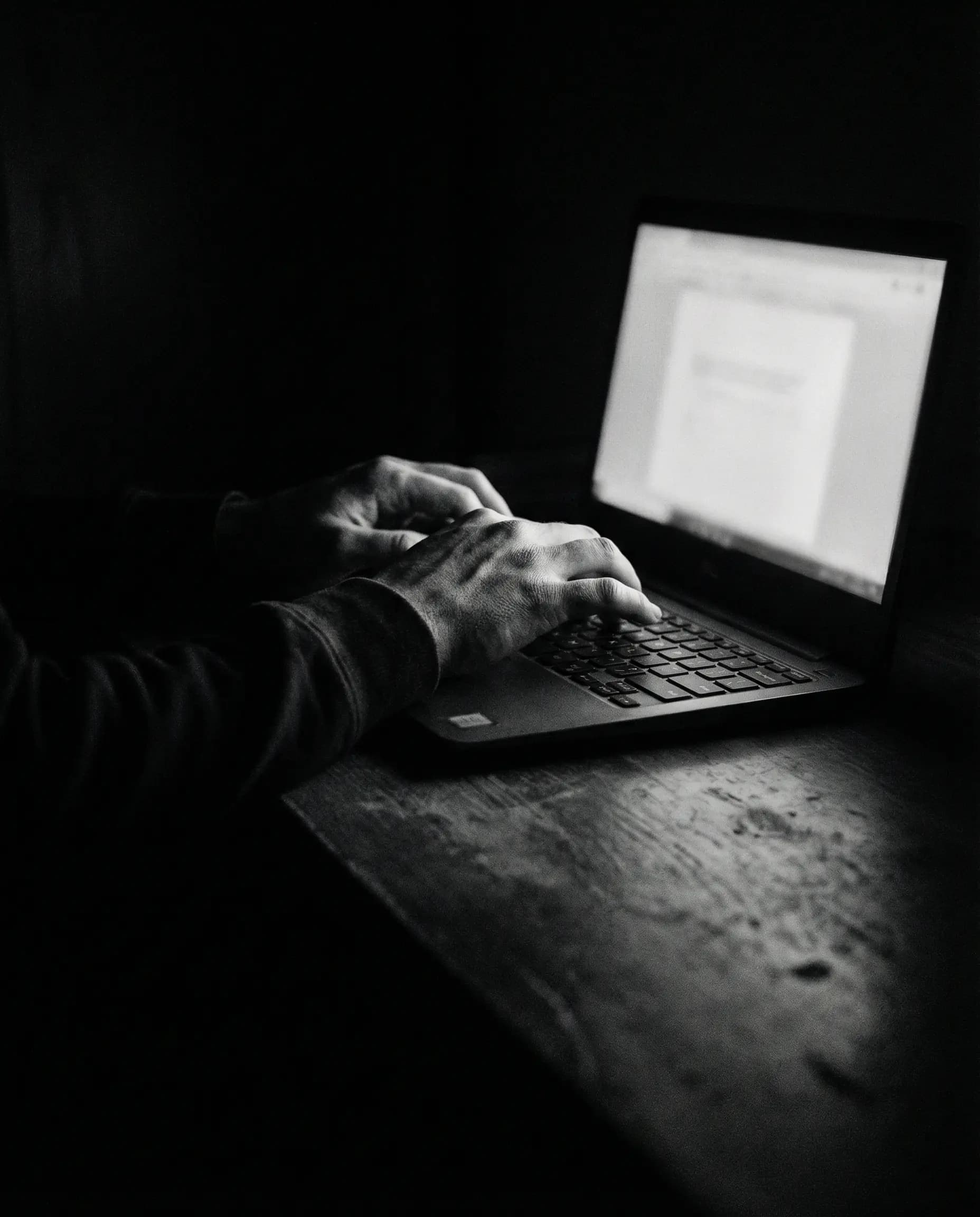 Hands typing on laptop in dark room with screen glow